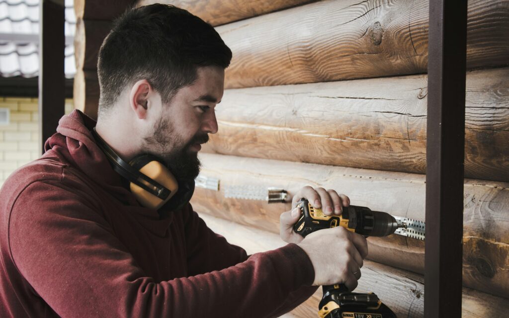 Satisfied worker is drilling wall. Builder is working construction site. Wooden house